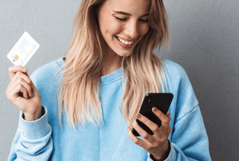 A woman in a blue sweater smiles while holding her phone and credit card. 