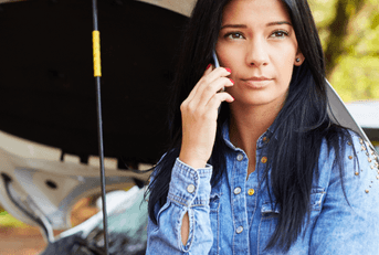 A woman leans against the open hood of her car while holding her phone to her ear.
