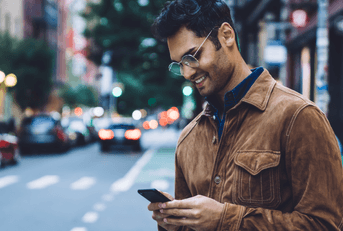 A man wearing glasses stands on the sidewalk in a busy city street while smiling and staring down at his phone.