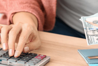 A woman in a pink cardigan uses a calculator on her desk while holding a stack of cash in front of her wallet.
