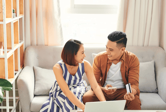 A couple sits on the couch together as they use the laptop in front of them and smile at one another.