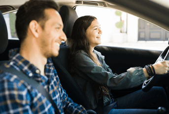 A woman smiles while driving her car, as a man smiles in her passenger seat.