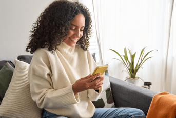 A woman in a creme colored sweater uses her phone while sitting on a gray couch.