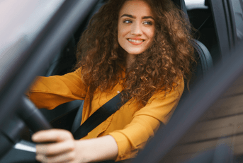A woman with curly hair and an orange shirt grips the steering wheel and smiles.