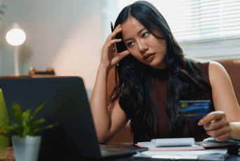 A woman holds a credit card and grips her head in frustration as she looks at bills on her computer. 