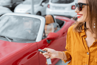 A woman wearing sunglasses uses her key fob to lock her red convertible.