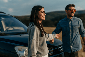 A couple stands in front of their vehicle while enjoying a mountain view.