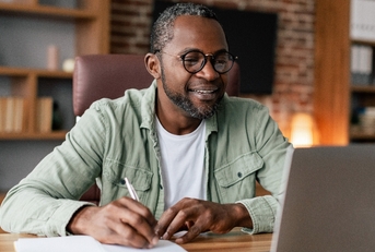 Man taking notes sitting at the desk looking at laptop