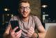 A man wearing glasses sits in front of a laptop and holding a smartphone. 