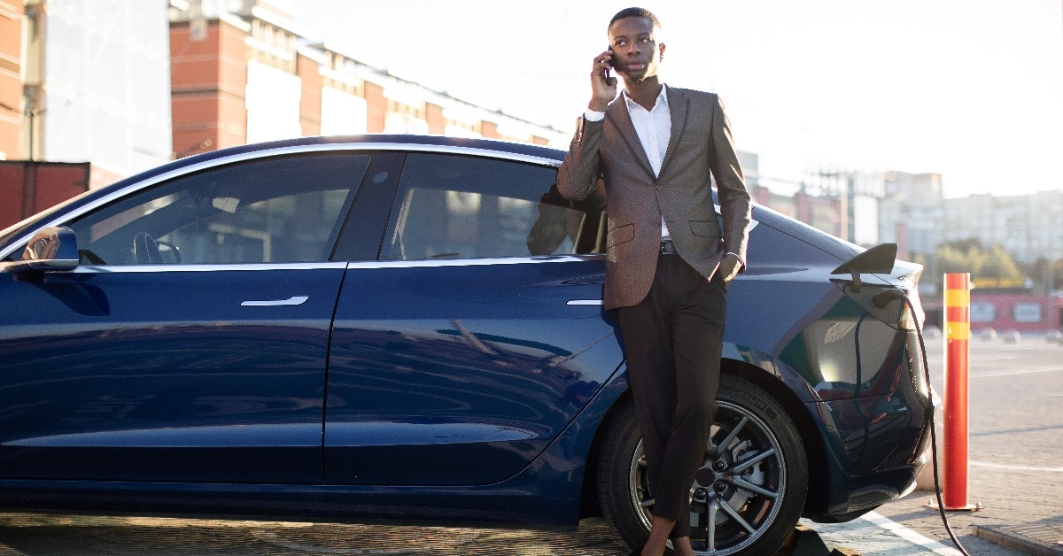  dark skinned man standing at the charging station beside his car