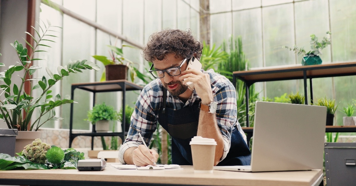 businessman talking on cellphone while standing in apron in small floral center