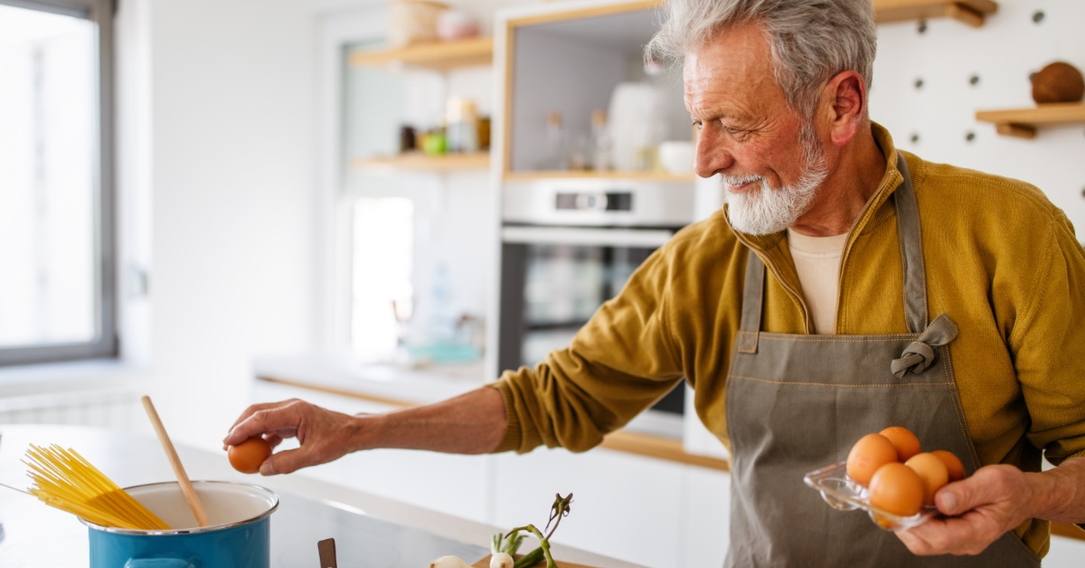 happy senior man cracking eggs and cooking pasta in kitchen