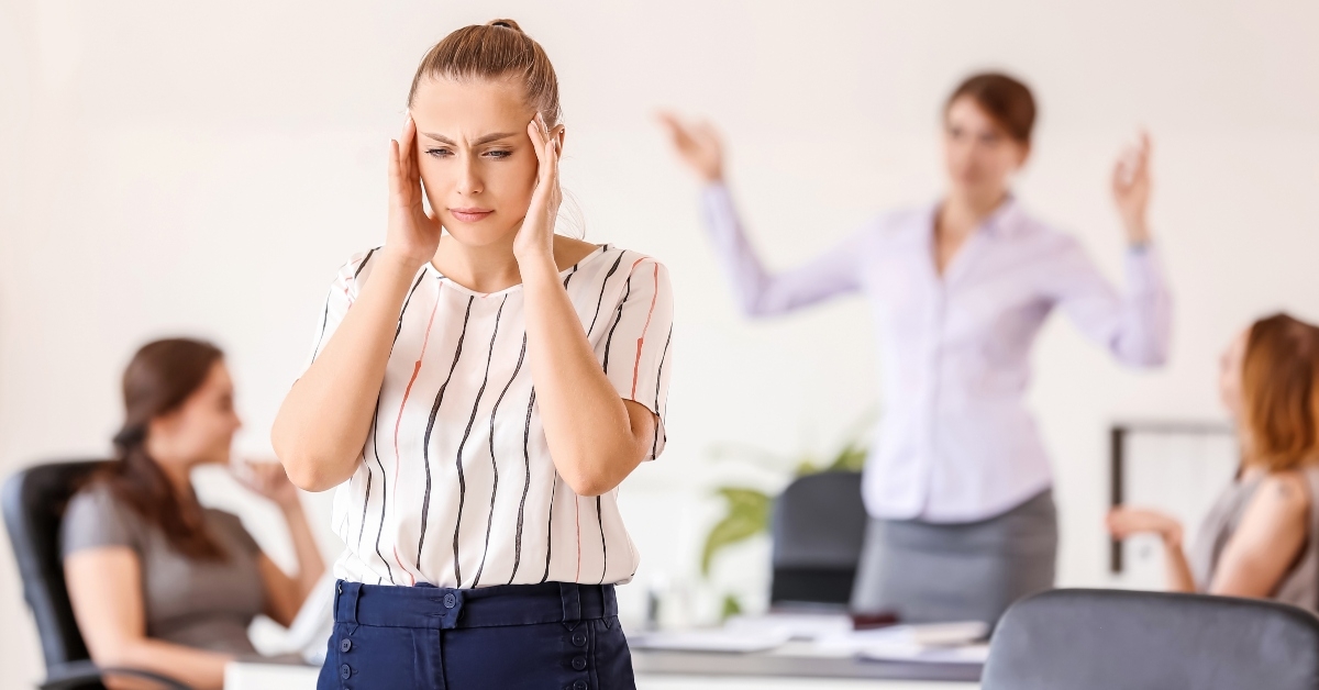Woman with hands on her head seems bothered while meeting in the back where other woman stands and talks with hands up