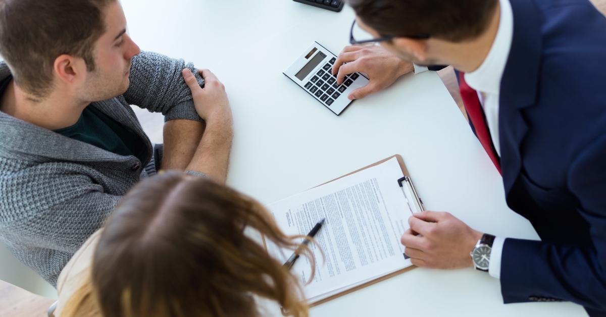 Couple looking at paper forms and calculator on the table being used by man in suit on the other side of the table