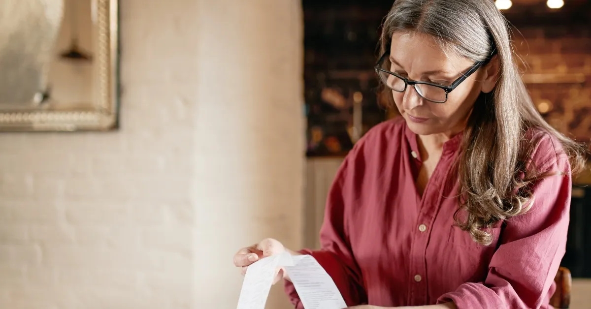senior woman on table with bills and cup focusing on long receipt 