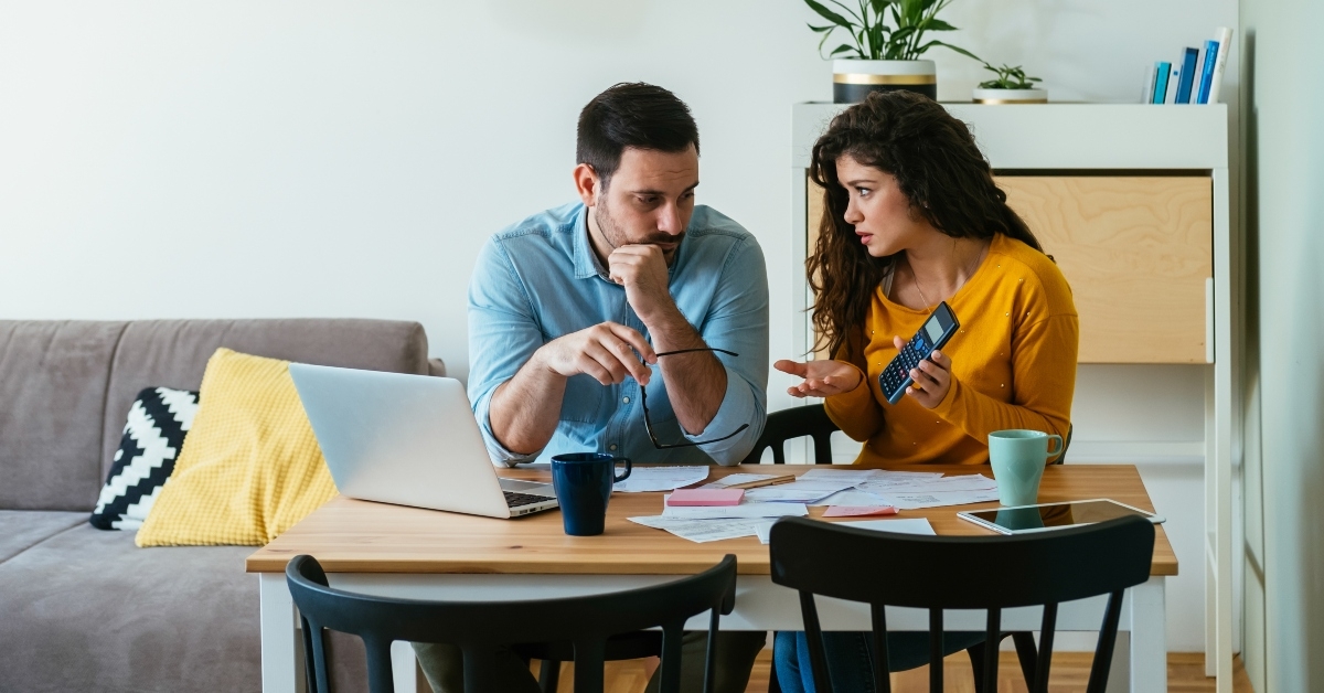 Woman shows man result in calculator both worried faces as they sit at living room table with papers, bills, laptop, and two mugs