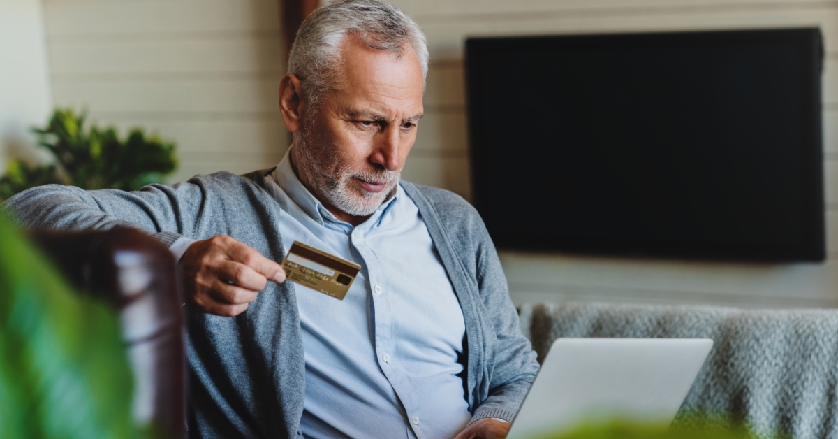 senior man sitting on couch at home working on laptop while holding credit card