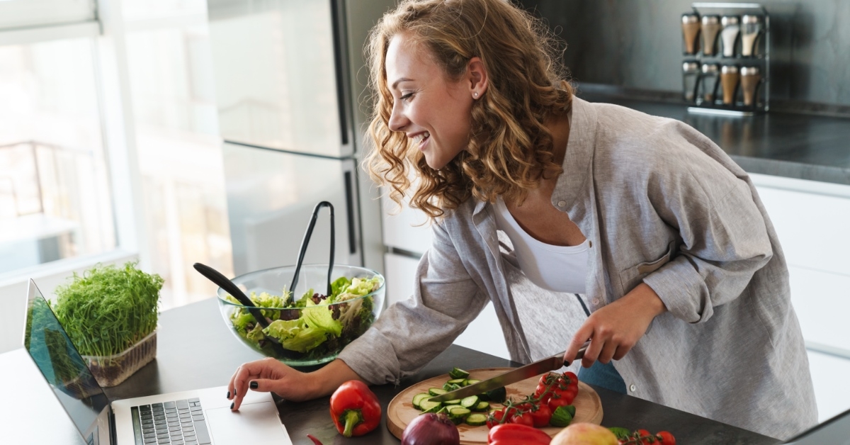woman standing in kitchen chopping vegetables while checking laptop