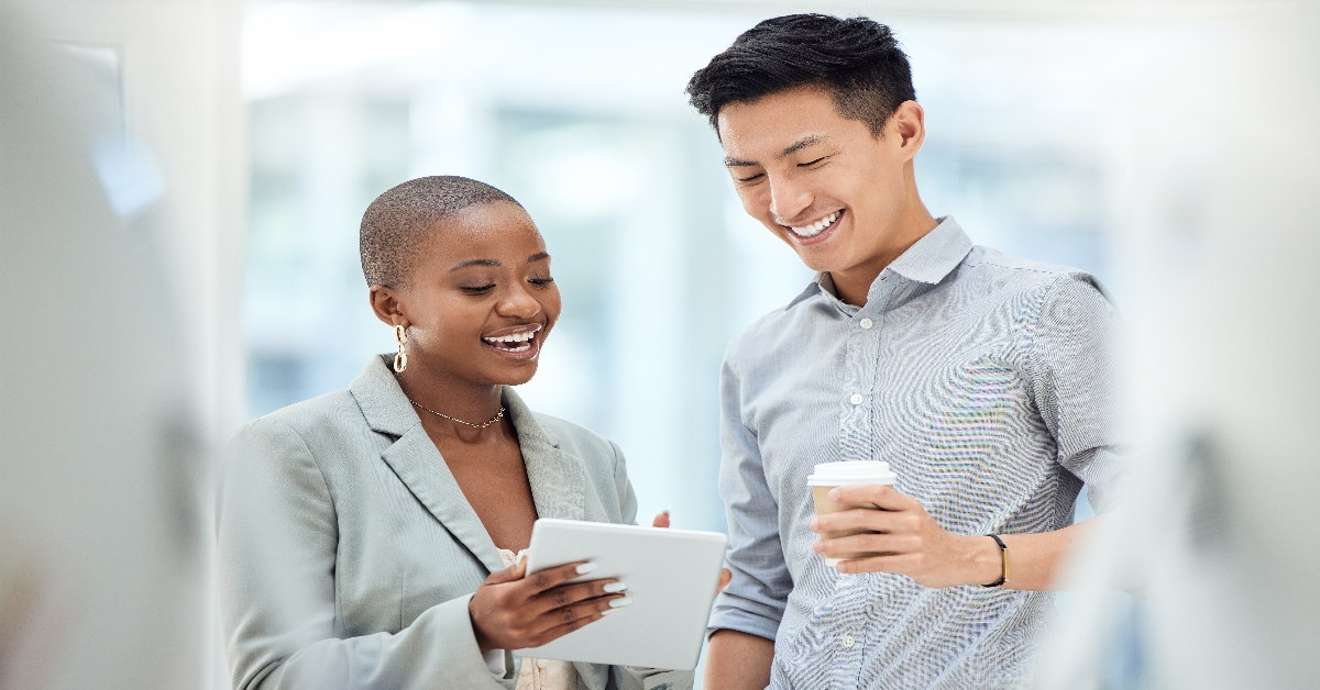 colleagues standing at workplace looking at tablet while holding coffee 
