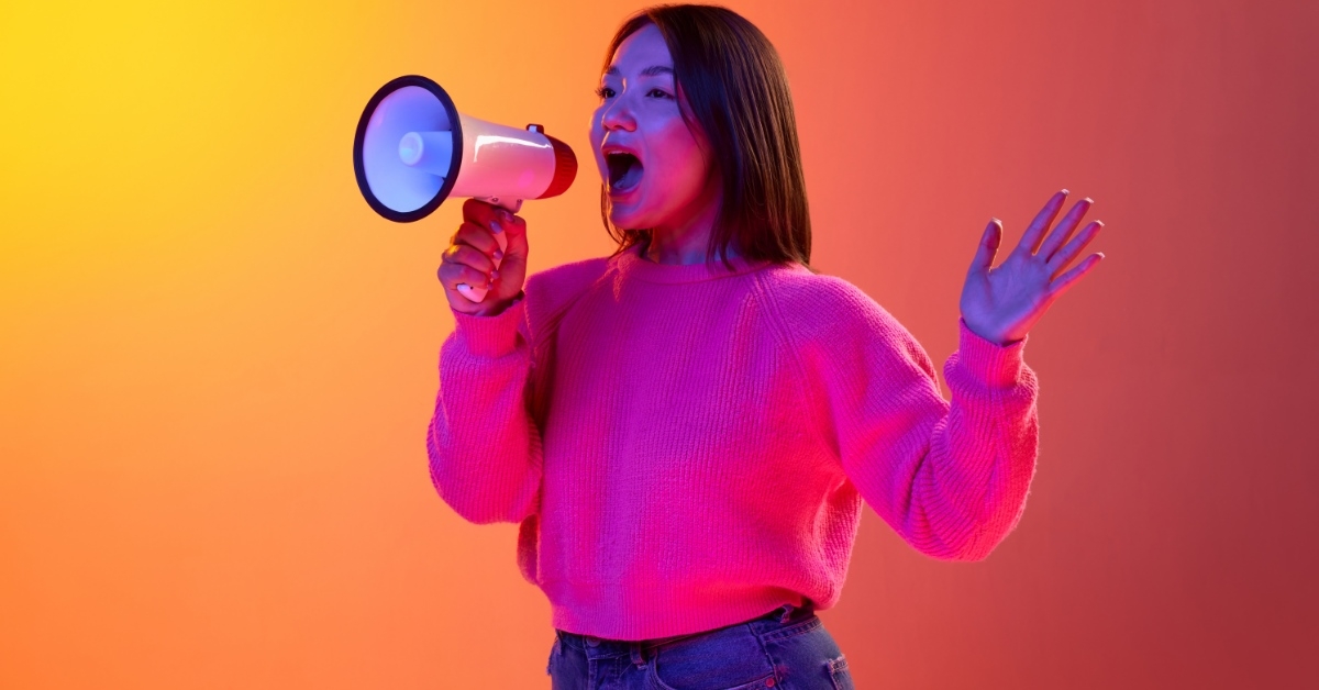 Woman in pink sweater talking on megaphone with neon orange background in back