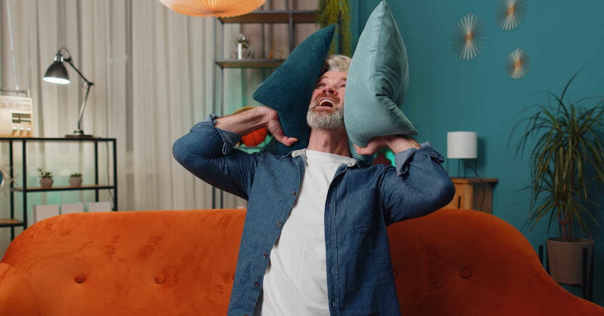 man sitting in couch at home covering his ears with cushions at home