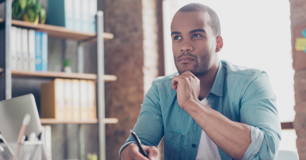 African American man sitting at table thinking about something while writing on notebook