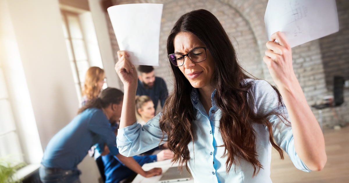 angry businesswoman holding papers in hands squinting eyes with employees talking in background