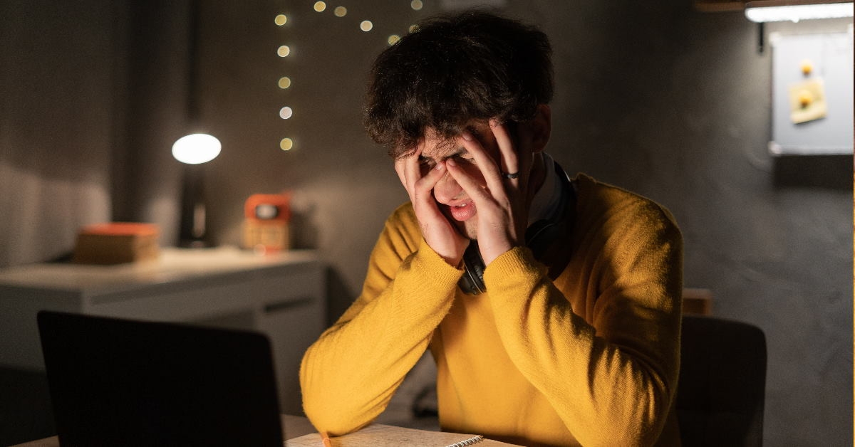 male student sitting in front of laptop on table covering face 