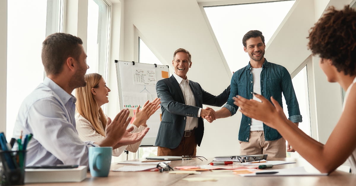 male boss shaking hand with male employee while colleagues are applauding in background