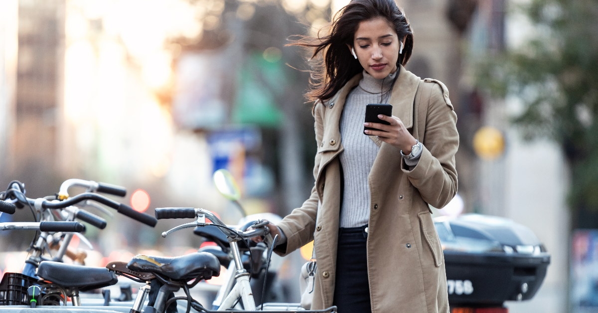 woman in long coat standing in street holding bicycle while using smartphone