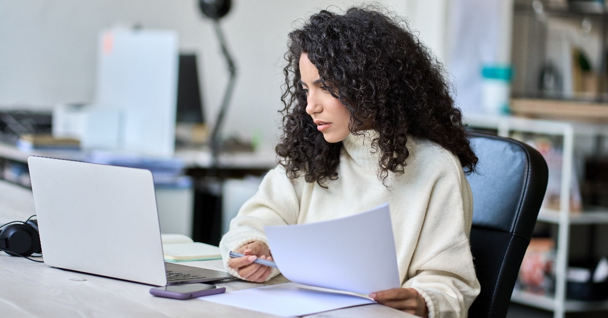 woman sitting in front of laptop at table reviewing documents in hand