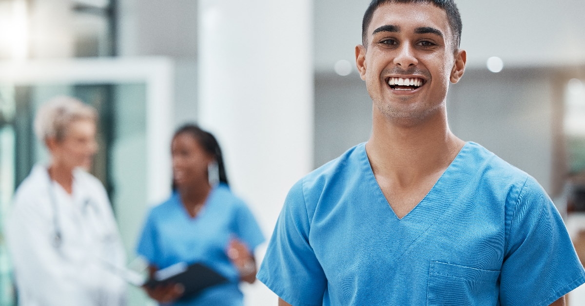 male healthcare nurse smiling while holding tablet at clinic with other colleagues in background