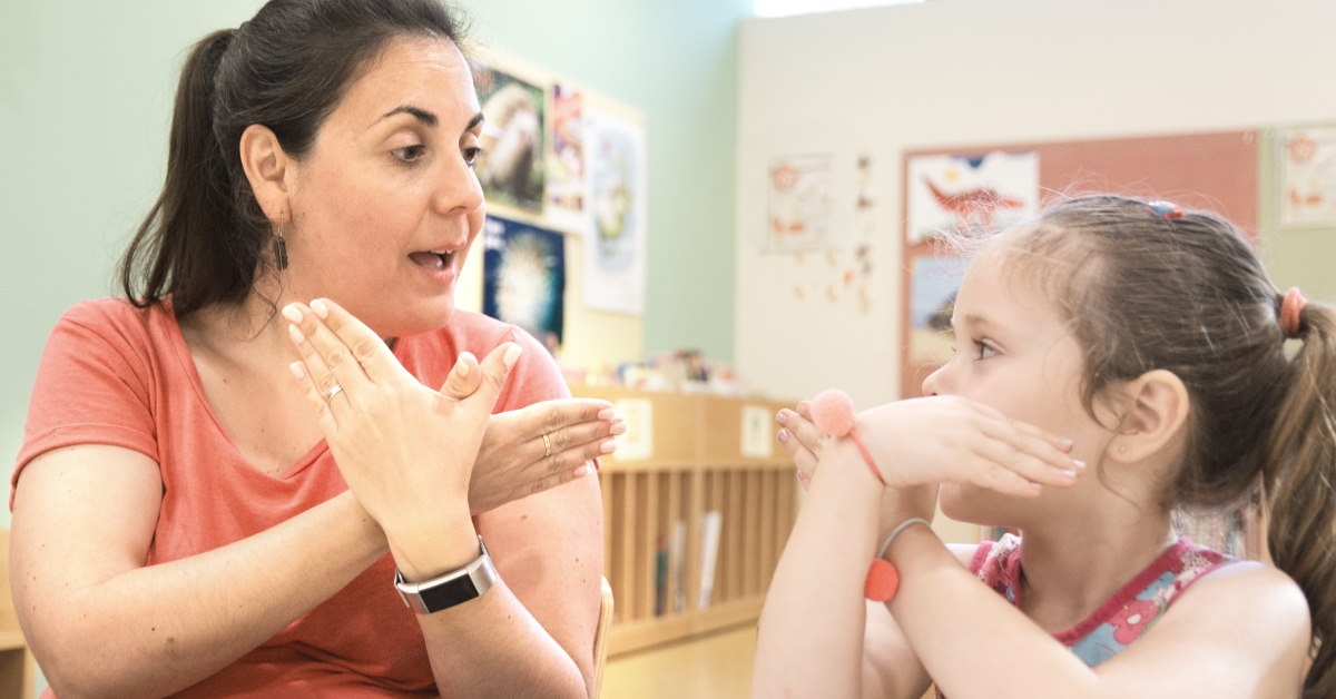 female tutor sitting in classroom teaching sign language to deaf girl 