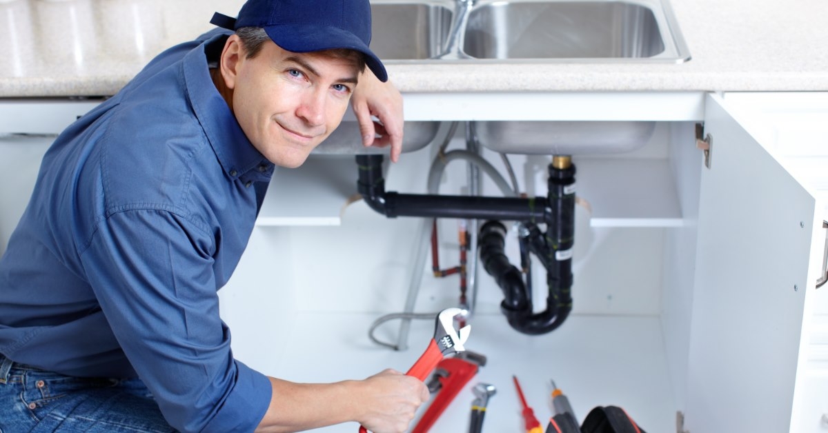 male plumber sitting on ground fixing kitchen sink holding wrench
