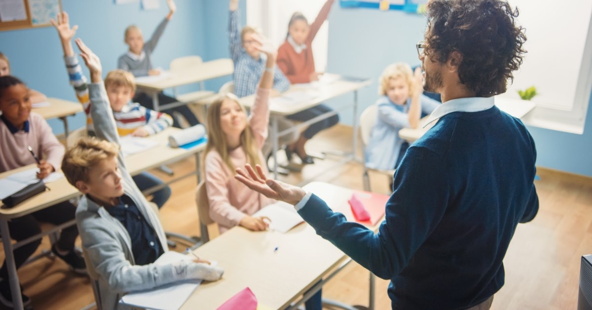 male teacher standing in elementary classroom with students raising hands and interacting with him