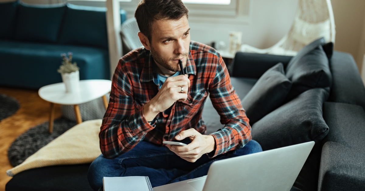 man thinking about finances while working on laptop and notebook on couch at home