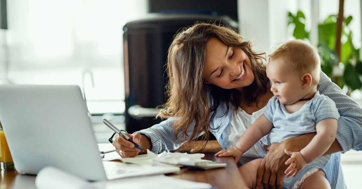 mother sitting at table with laptop writing on paper while talking to baby at home