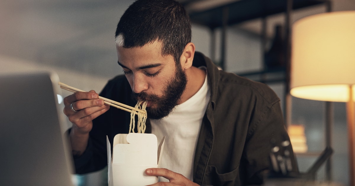 man using chopsticks to eat takeaway ramen while sitting in office