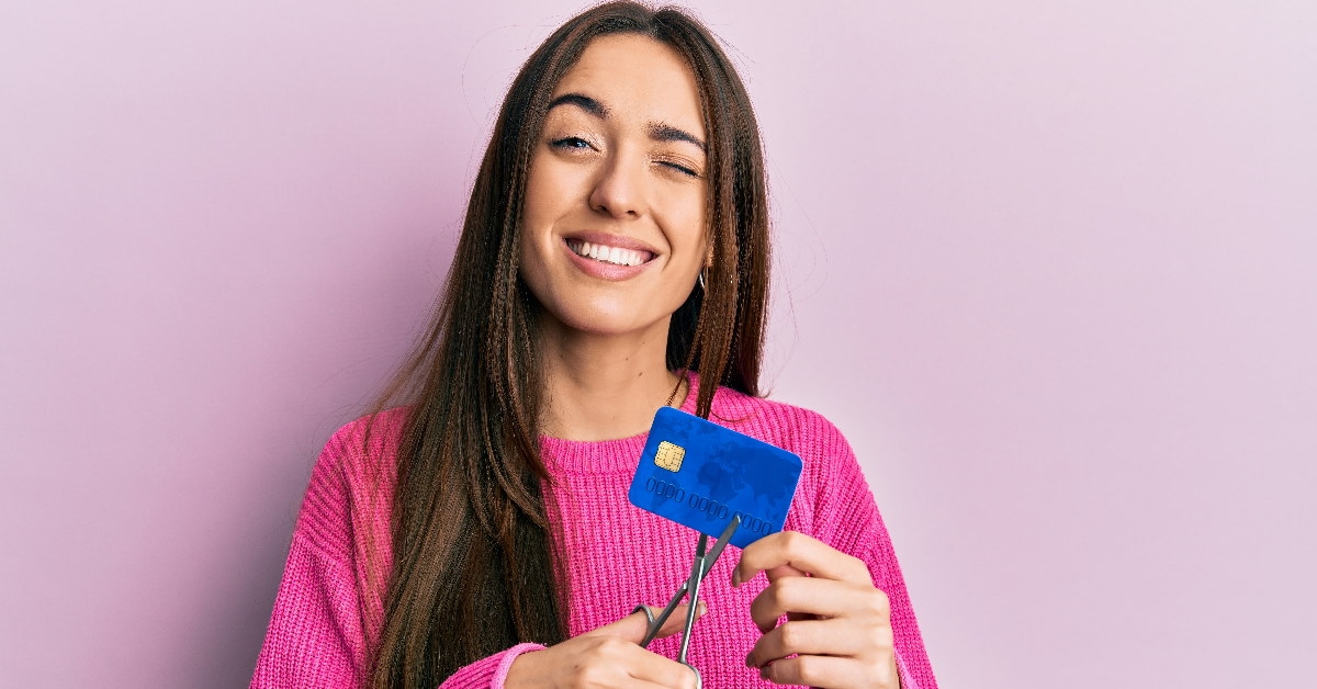 girl in hot pink sweater winking while cutting credit card with scissors standing on baby pink background 