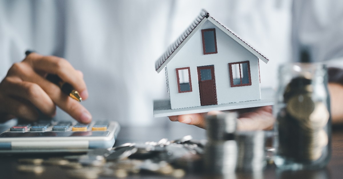 man holding home sculpture in hand while calculating money for investment on calculator and pennies on table