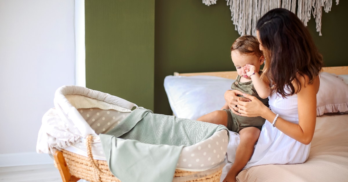 mother holding baby boy in lap besides bassinet sitting on bed