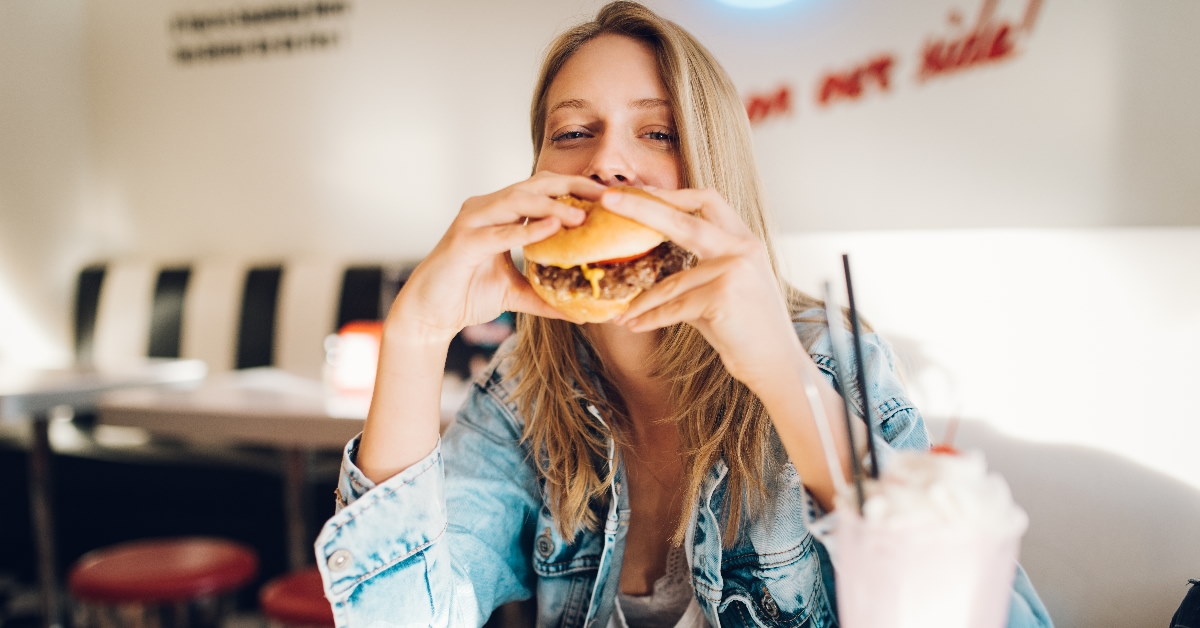 woman sitting in restaurant with shake and fries on white table eating a burger in hand