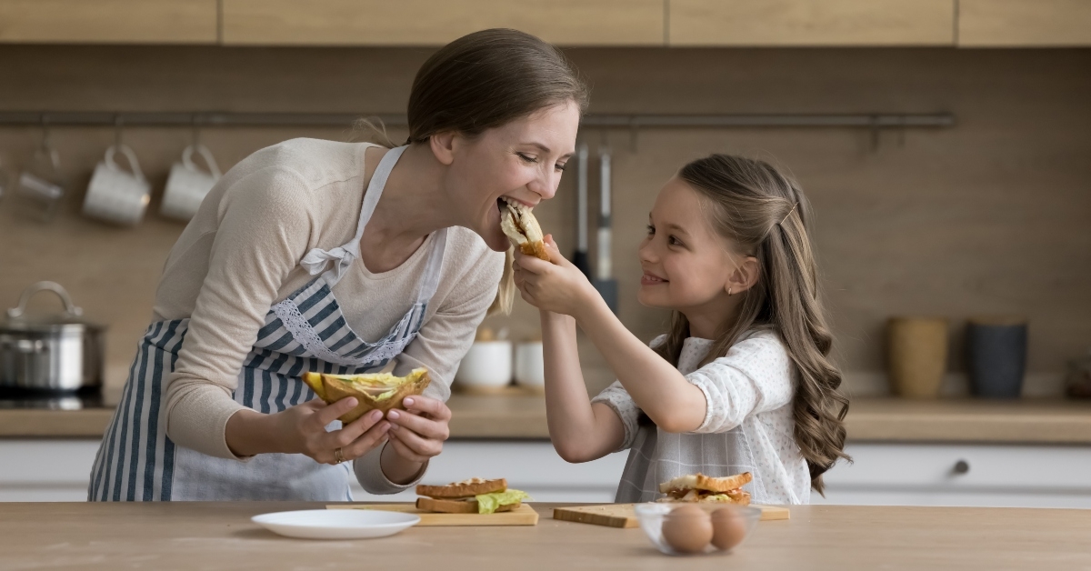 joyful little kid girl giving mom sandwich to bite