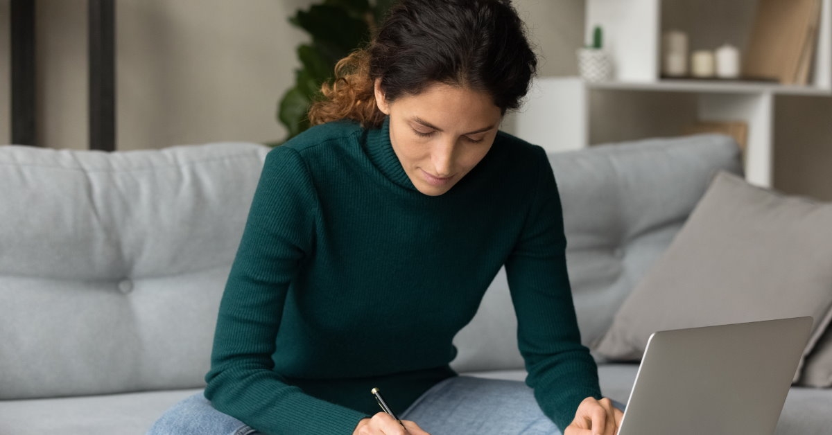 woman sitting on couch in front of laptop writing family budget on notebook
