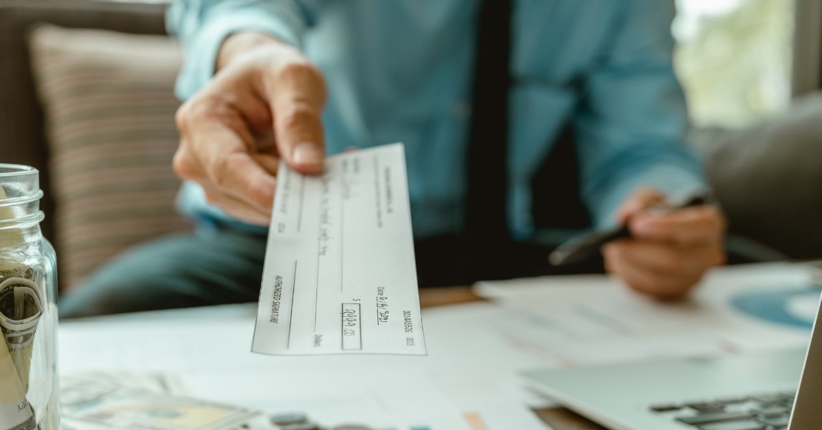 businessman in formal shirt handing over cheque to someone with pennies and money on table beside laptop