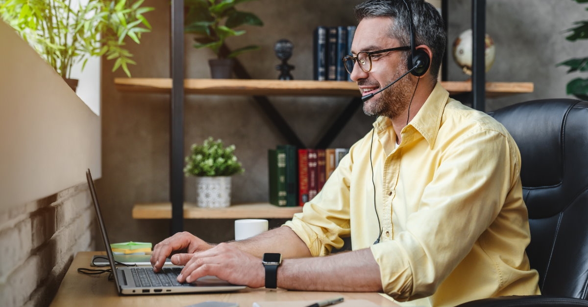 senior aged man sitting at his work desk using laptop while wearing a headset