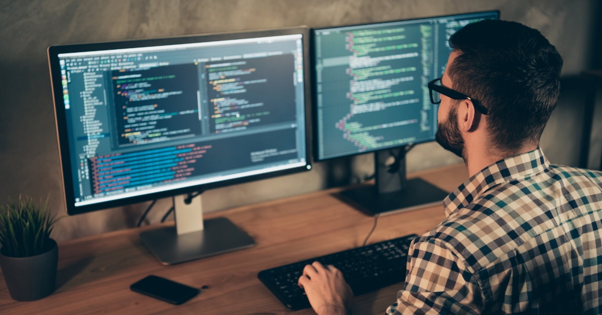 young man sitting at wooden table coding on two monitors