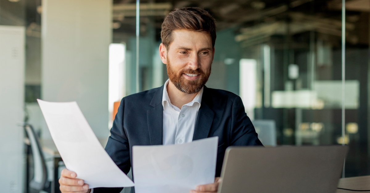 businessman sitting at office table reviewing documents while working on laptop