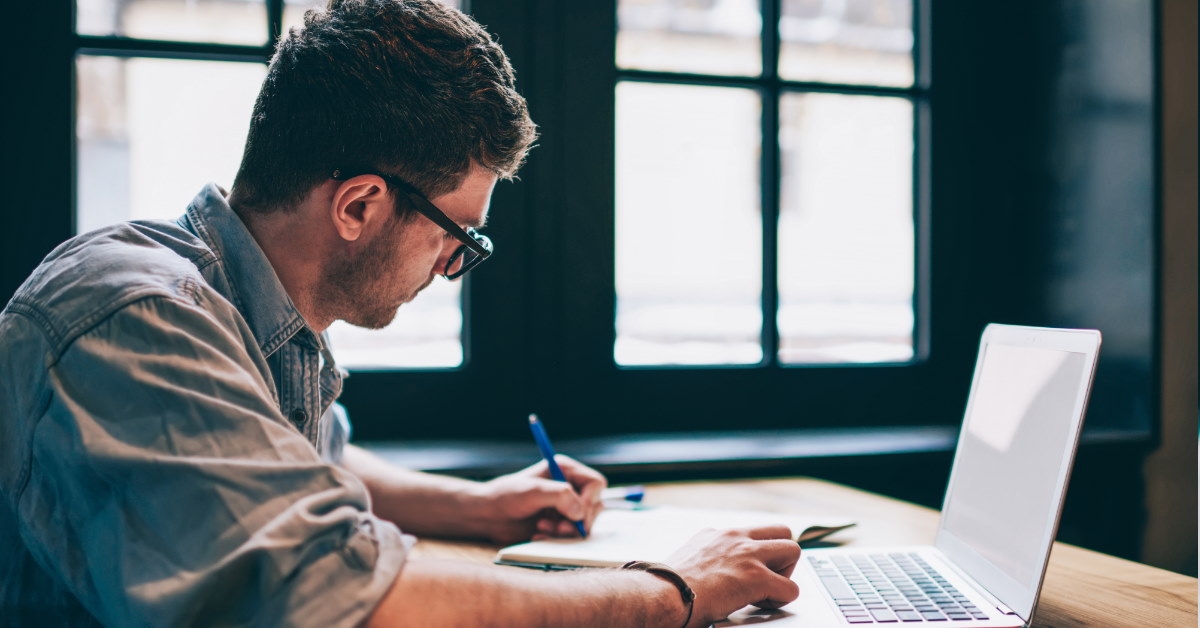 male student sitting at table using laptop to prepare academic notes