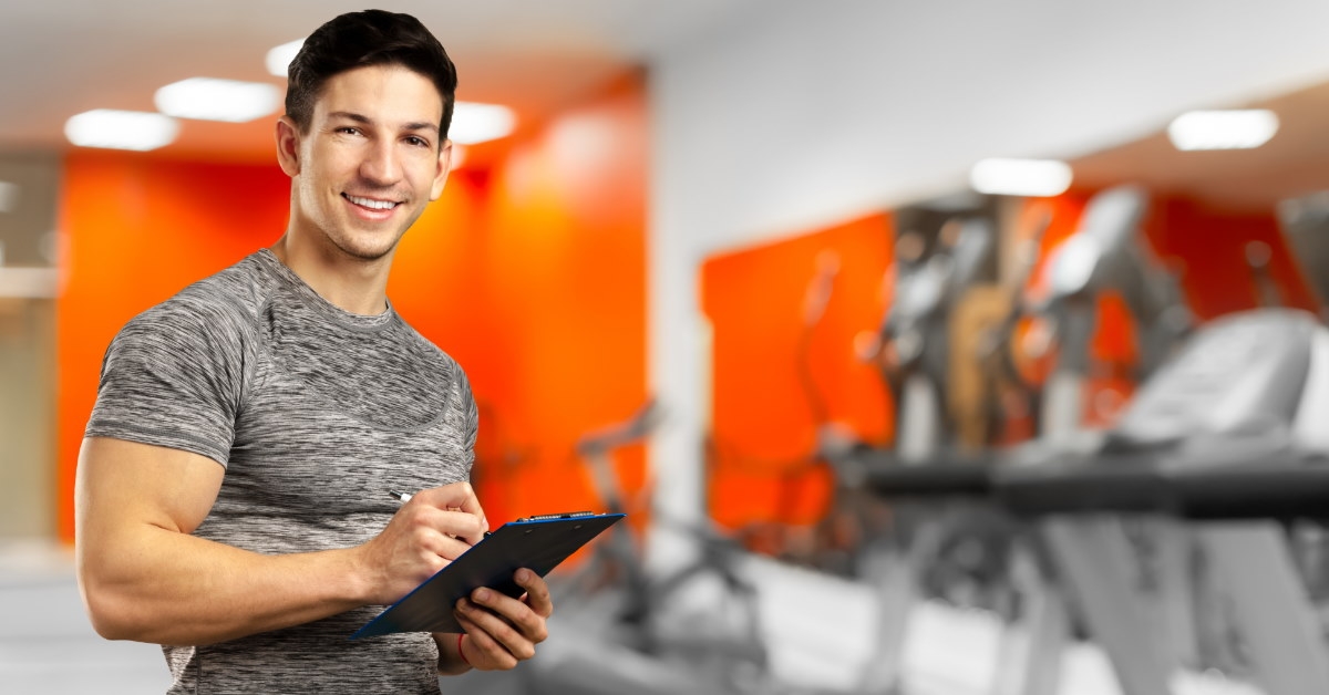 male personal trainer standing in gym with treadmills at back writing on notepad 